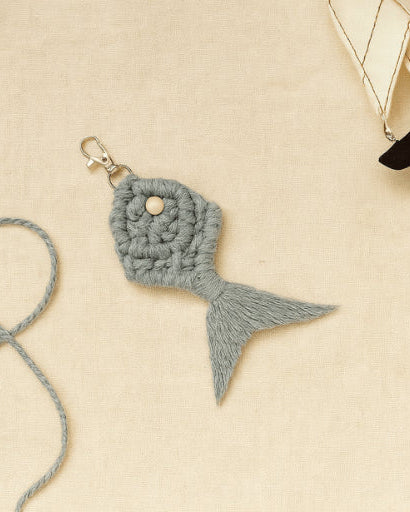 Handmade macramé fish keychain in grey-blue, styled with nautical props and cotton string on a light beige linen background