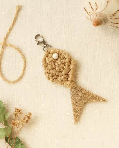 Boho-style macramé fish keychain in light brown, surrounded by eucalyptus leaves, dried flowers, and a decorative shell on a soft fabric backdrop