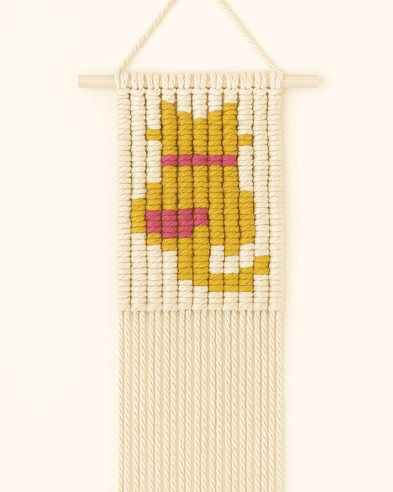 A rectangular macramé wall hanging features a mustard yellow cat with a pink heart collar in a pixel-style design, knotted with off-white cotton cord and hanging on a wooden dowel.