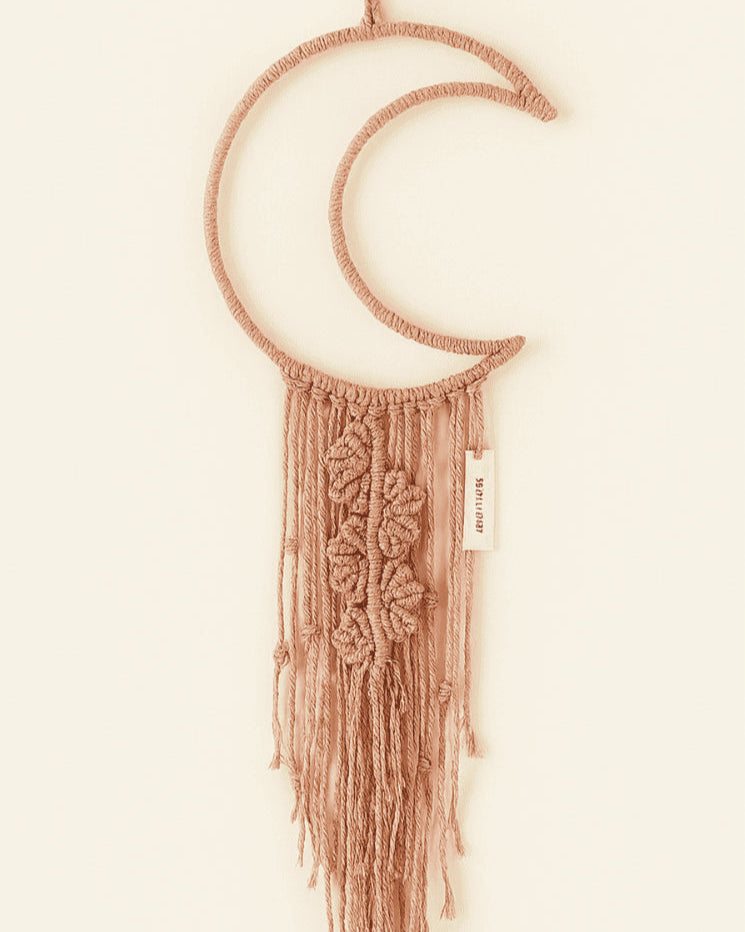 Boho macramé wall hanging shaped like a crescent moon in dusty pink, featuring floral-inspired knots and long fringe. Neutral backdrop, perfect for cozy home decor.