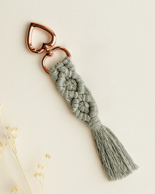 Grey handmade macramé diamond keychain with rose gold heart clasp, styled on an off-white background with dried florals