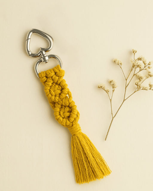 Handmade macramé diamond keychain in mustard yellow with silver heart clasp, styled on an off-white background with dried flowers