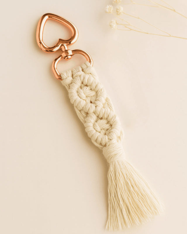 Off-white handmade macramé keychain with rose gold heart clasp, laid on a soft neutral background with dried baby's breath