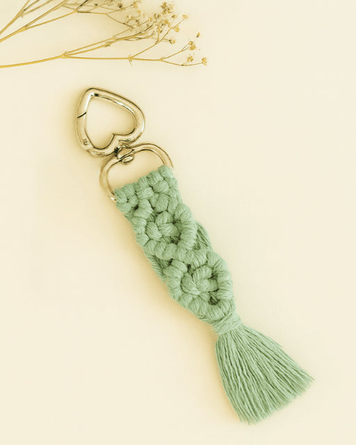 Soft sage green macramé keychain with silver heart clasp, displayed on a neutral background with a sprig of dried flowers