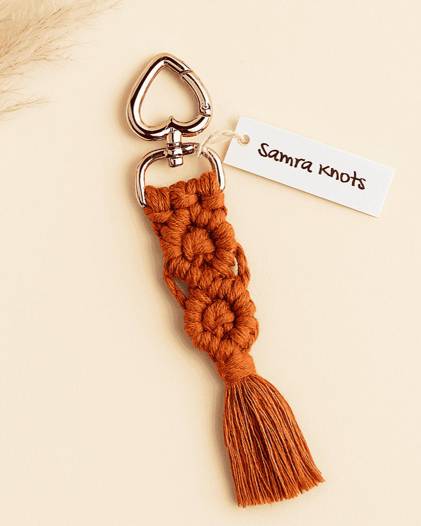 Terracotta handmade macramé keychain with rose gold heart clasp, styled with neutral pampas grass on an off-white background
