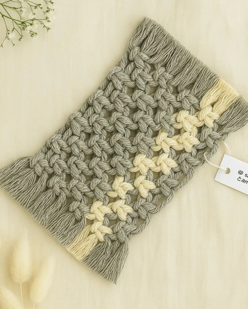 Two-tone macramé mug rug handmade by Samra Knots in grey and off-white, displayed on a soft beige linen background with fluffy dried florals and a product tag for branding.