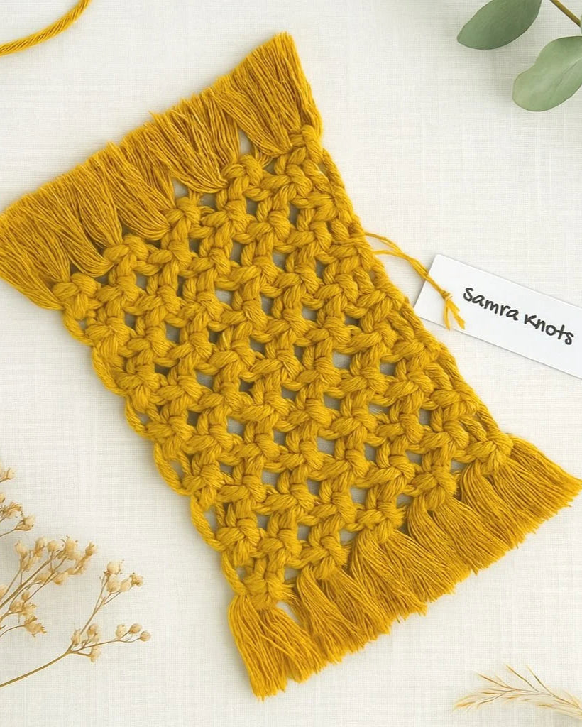 Flat lay of a handcrafted mustard yellow macramé mug rug by Samra Knots, styled with eucalyptus leaves and dried florals on a soft beige fabric background.