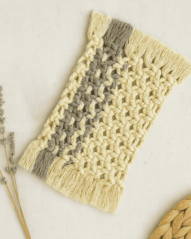 Boho-style macramé mug rug in off-white with a grey stripe, featuring fringed edges and a Samra Knots tag, styled with dried grass on a neutral fabric backdrop.