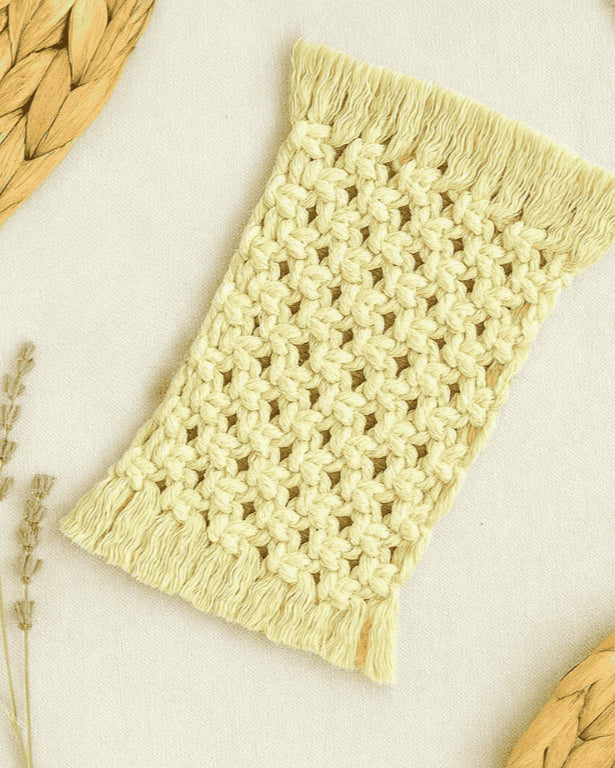 Flat lay of a handmade off white macramé mug rug by Samra Knots, styled with dried lavender sprigs and natural woven placemats on a neutral beige fabric background.
