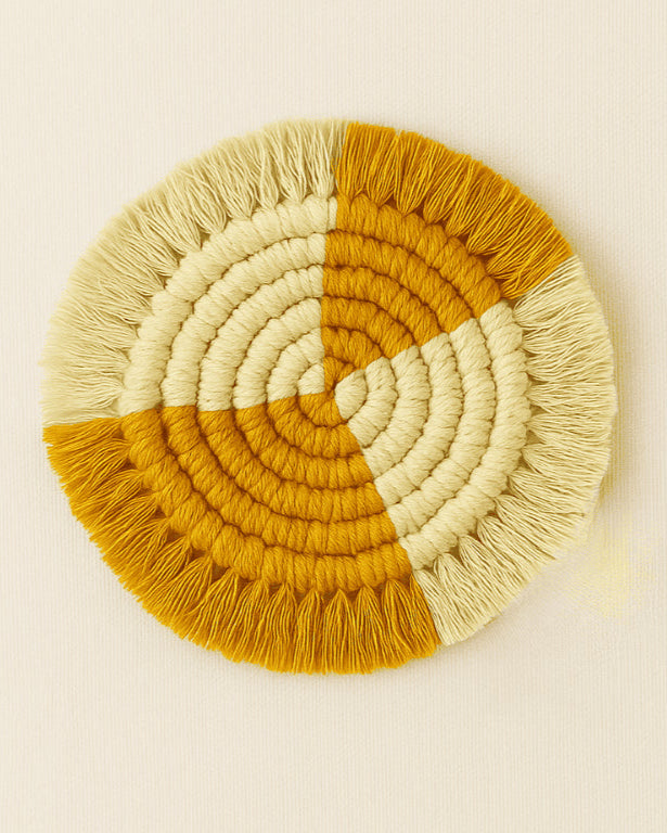 A handmade round macramé coaster featuring a quartered design with alternating muted mustard and natural cotton colors, displayed on a soft beige background.