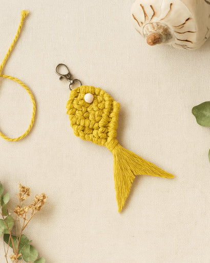 Handmade macramé fish keychain in mustard yellow, styled on a neutral fabric background with natural dried florals and cotton cord.