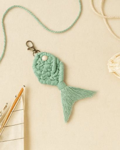 Boho-style sage green macramé fish keychain placed on a warm beige backdrop, surrounded by cotton twine and a mini sailboat decor.