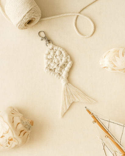 Soft off-white handmade macramé fish keychain styled with carved seashells, cotton spool, and a mini boat on a light neutral background.