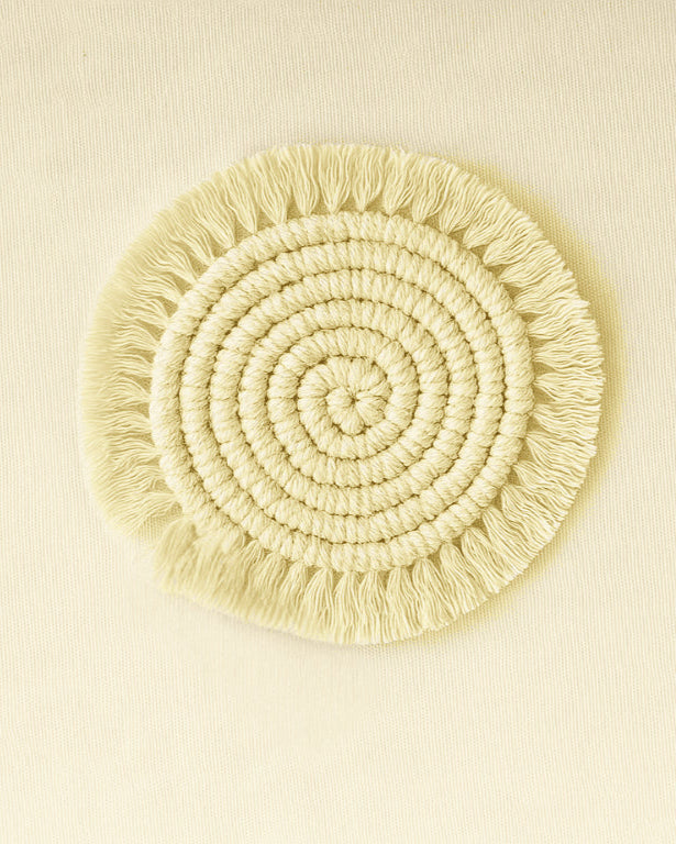 A soft, round macramé coaster made of natural cotton yarn with spiral knotting and fringe edges, set on a beige canvas .