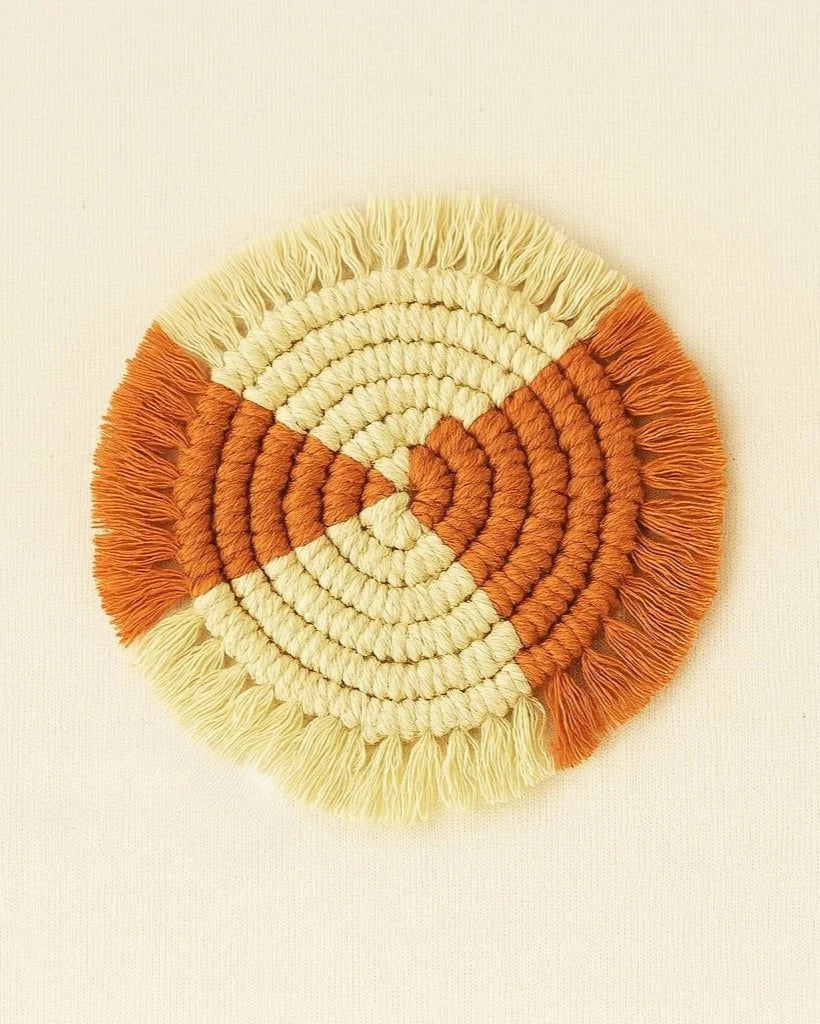 A handmade round macramé coaster featuring a quartered design with alternating terracotta and natural cotton colors, displayed on a soft beige background.