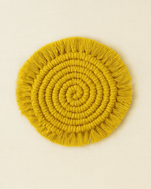 A circular macramé coaster in mustard yellow with tightly coiled knots and fringed edges, placed on a neutral beige background .