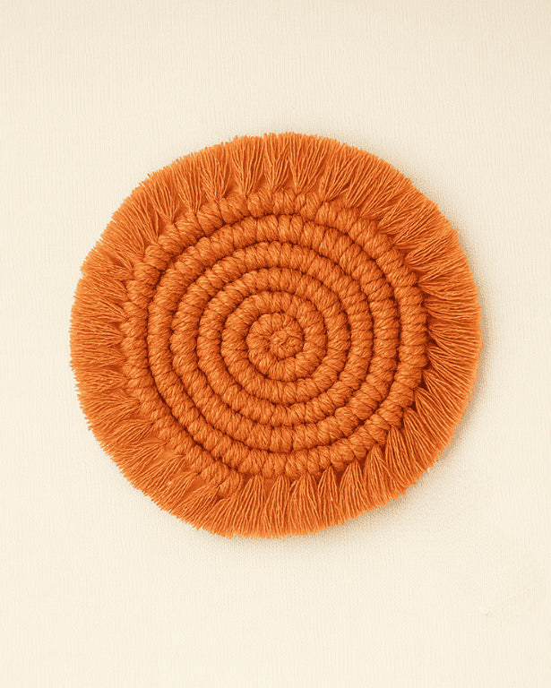 A handcrafted round macramé coaster in a warm terracotta color with a spiral design and fringe edges, lying flat on a beige textile background.