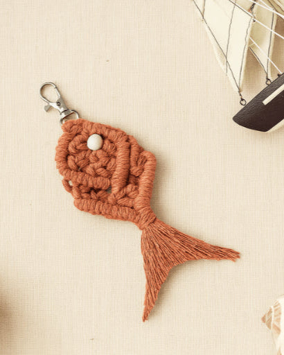 Earthy terracotta-colored macramé fish keychain laid flat on beige fabric with matching yarn, mini ship, and nautical-inspired styling.