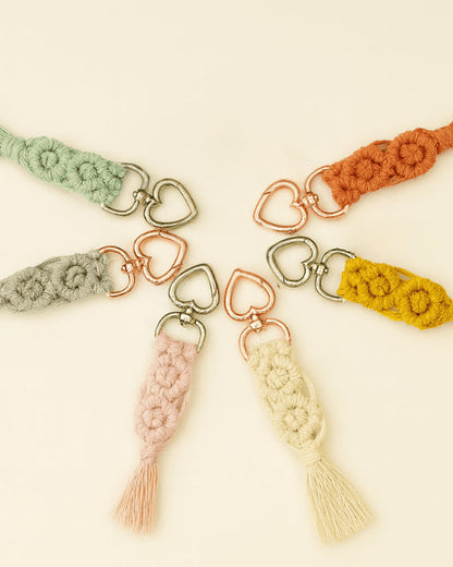 Flat lay of seven handmade macramé diamond keychains arranged in a circle, each in a different colour with heart-shaped metal clasps on a soft neutral background