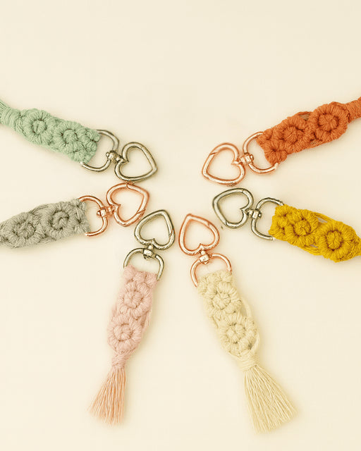 Flat lay of seven handmade macramé diamond keychains arranged in a circle, each in a different colour with heart-shaped metal clasps on a soft neutral background