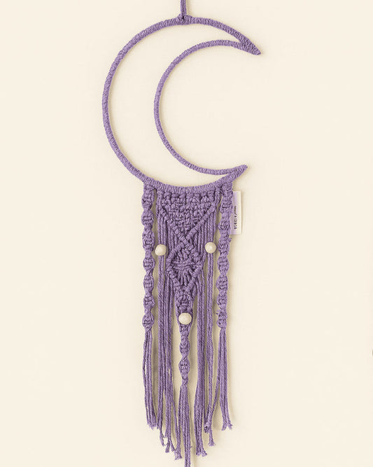 Handmade macramé crescent moon wall hanging in muted purple with natural wooden beads, displayed on a soft off-white background. Boho-style nursery or bedroom decor.