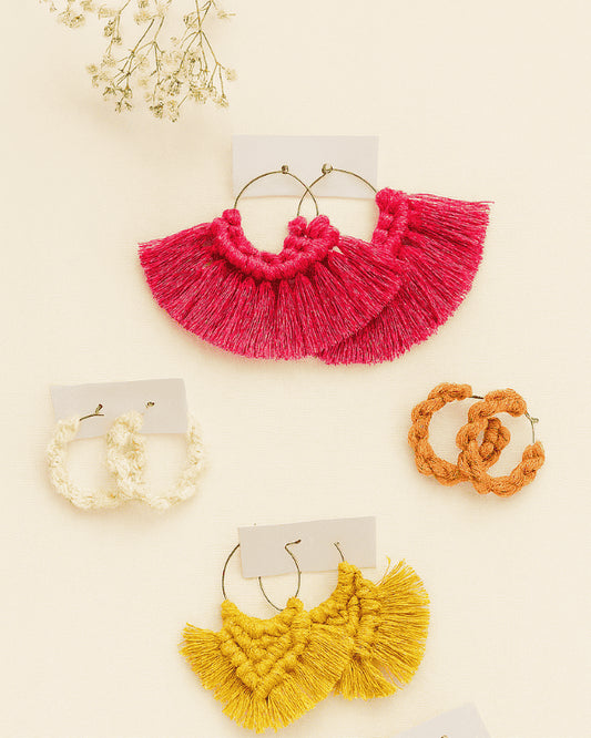 Four pairs of handmade macramé hoop earrings in pink, mustard yellow, terracotta, and off-white displayed on a neutral background with baby’s breath flowers