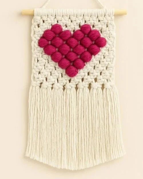 A handmade macramé wall hanging with a raised pink heart on an off-white cotton background and fringe, hanging from a wooden dowel.