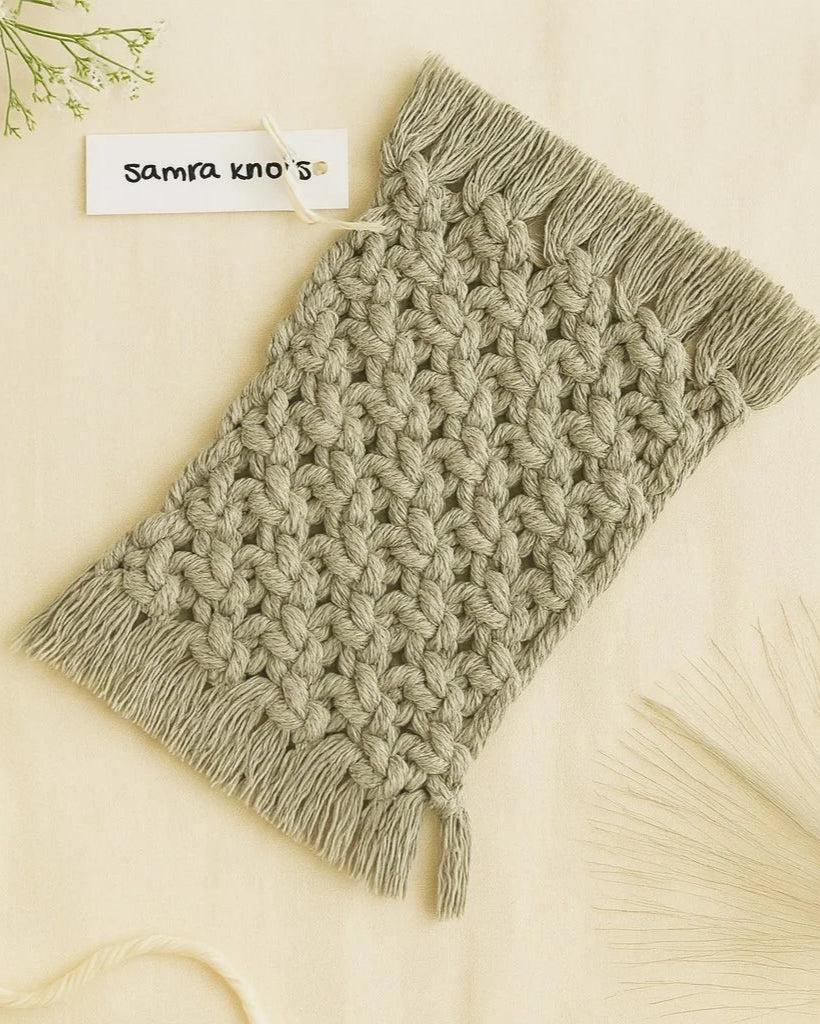 Handmade macramé mug rug in grey by Samra Knots, styled on a soft beige linen background with dried floral accents and a cotton rope for a natural boho aesthetic.