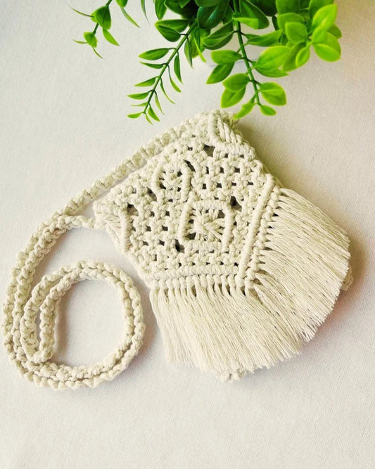 Handmade off-white macramé crossbody bag with fringe and intricate knot design, styled with green leaves – Samra Knots.