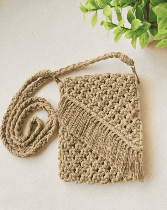 Full flat-lay of a light brown macramé crossbody bag with braided strap and fringe, displayed with green plant decor on neutral background.