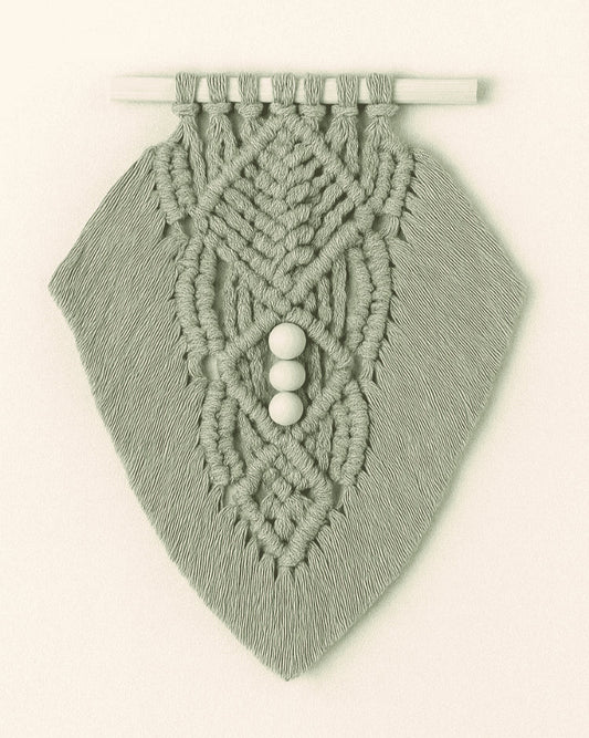 Boho macramé wall hanging in muted sage green, featuring leaf-inspired knots and three wooden beads, photographed on neutral backdrop.