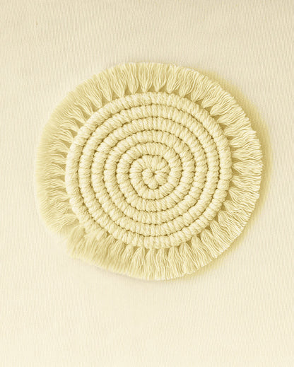 A soft, round macramé coaster made of natural cotton yarn with spiral knotting and fringe edges, set on a beige canvas .