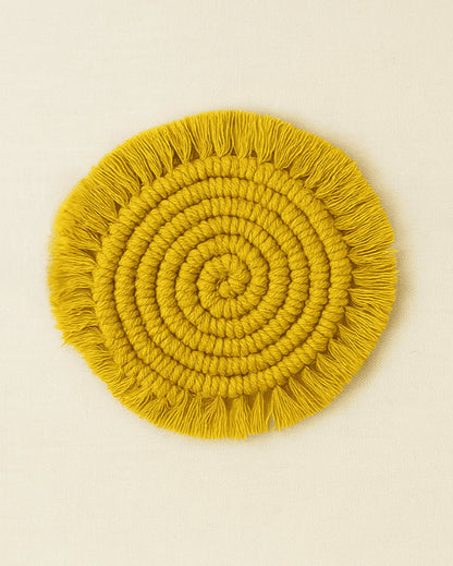 A circular macramé coaster in mustard yellow with tightly coiled knots and fringed edges, placed on a neutral beige background .