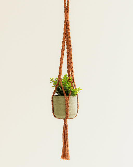 Handmade macramé plant hanger in terracotta, hanging on a light background with a concrete pot and green plant – boho home décor style