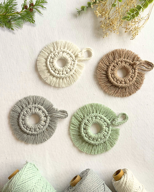 Four handmade macramé mini wreath ornaments in neutral tones, displayed on an off-white background with soft greenery and yarn spools.